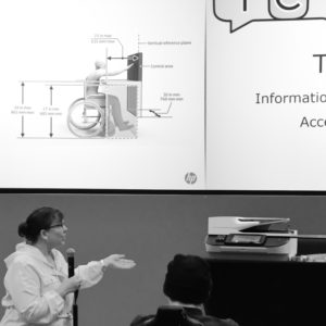 Laura Renfro presenting on hardware testing with a desktop printer on hand. Slide shows wheelchair reach range diagram.